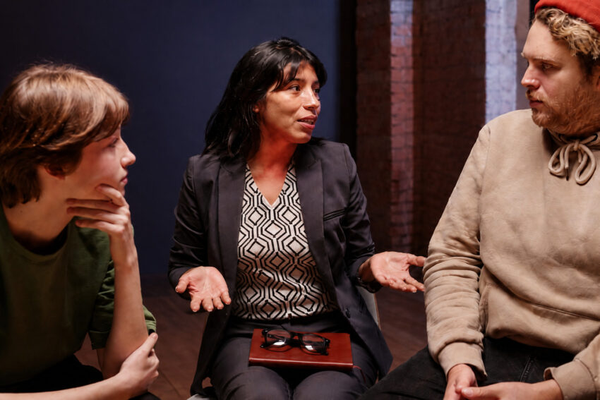 Drie personen zitten in een kring en voeren een open en rustig gesprek, waarbij de vrouw in het midden iets uitlegt met open handgebaren.